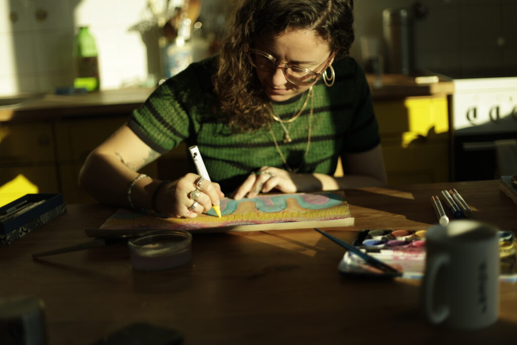 A white woman with long dark hair and glasses is drawing with chalk markers. She is wearing a green top, gold necklaces, hoop earings, and has many rings on.