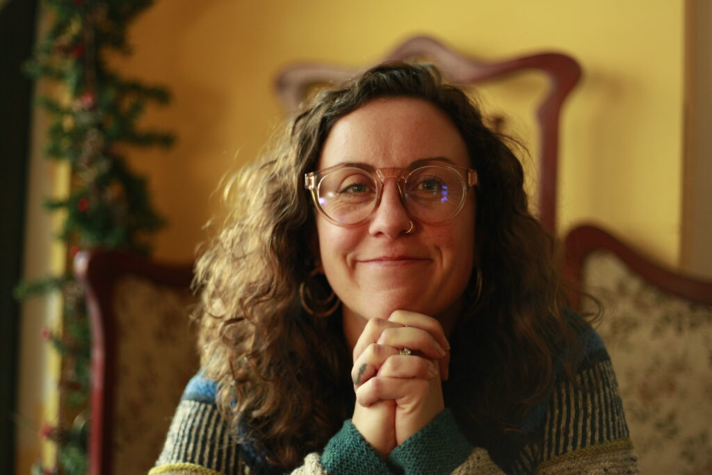 A white woman is looking directly into the camera and smiling. She has big, dark curly hair. She is wearing glasses. She is resting her chin on her folded hands.