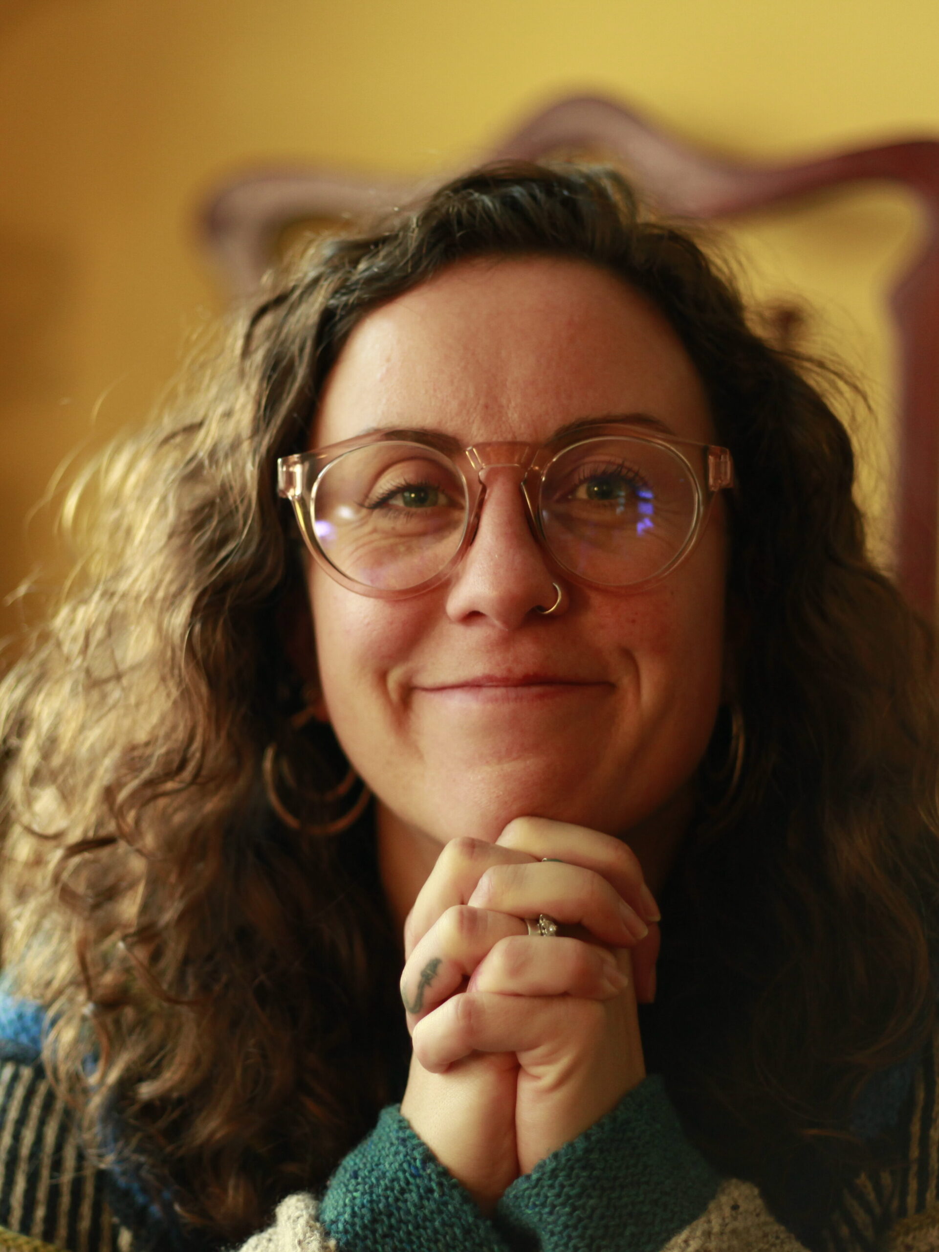 Alice is a white woman with big curly brown hair. She is smiling and resting her chin on her hands. She's wearing a blue, green, and stone grey jumper.