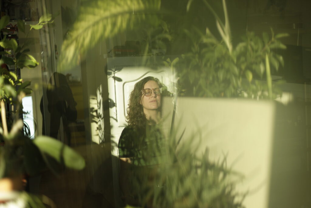 A white woman with glasses on is meditating. There are green plants in the foreground. She is sitting in a beam of sunlight.