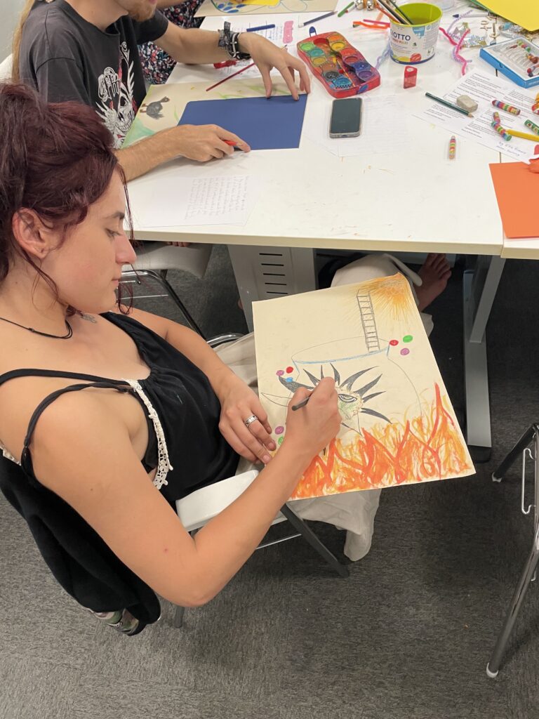 A woman is sitting at a chair. She has a piece of paper on her lap and is drawing. She looks relaxed and happy.